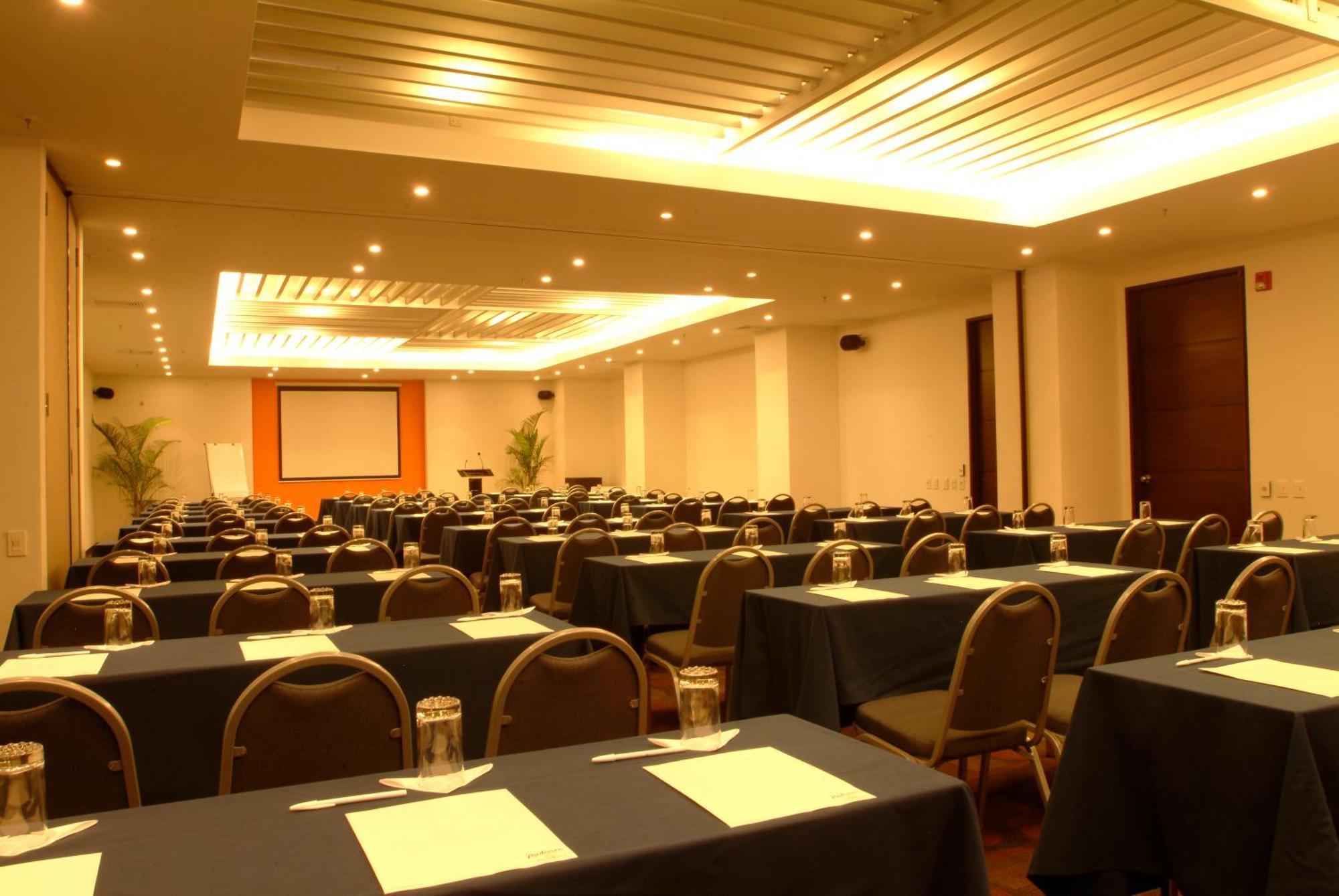 Decapolis Hotel Panama City Exterior photo The photo shows a conference or meeting room set up for an event. There are multiple rows of tables arranged with dark-colored tablecloths. Each table is equipped with a piece of paper or a notepad, and there are glass jars or cups placed on the tabl